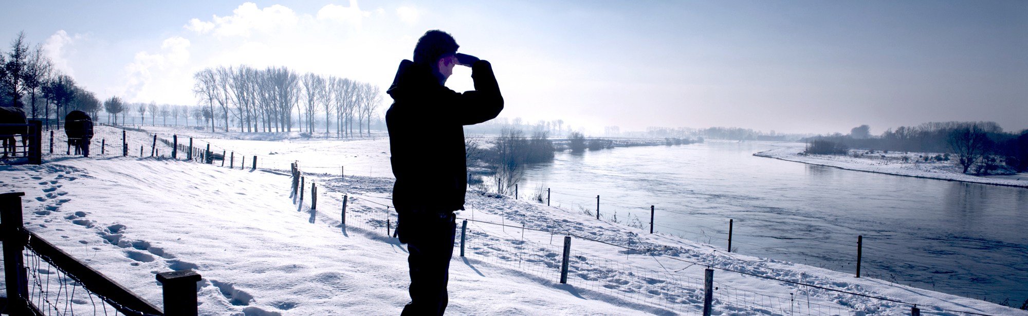 Wandelaar die uitkijkt over de Maas vanaf een winters besneeuwde oever