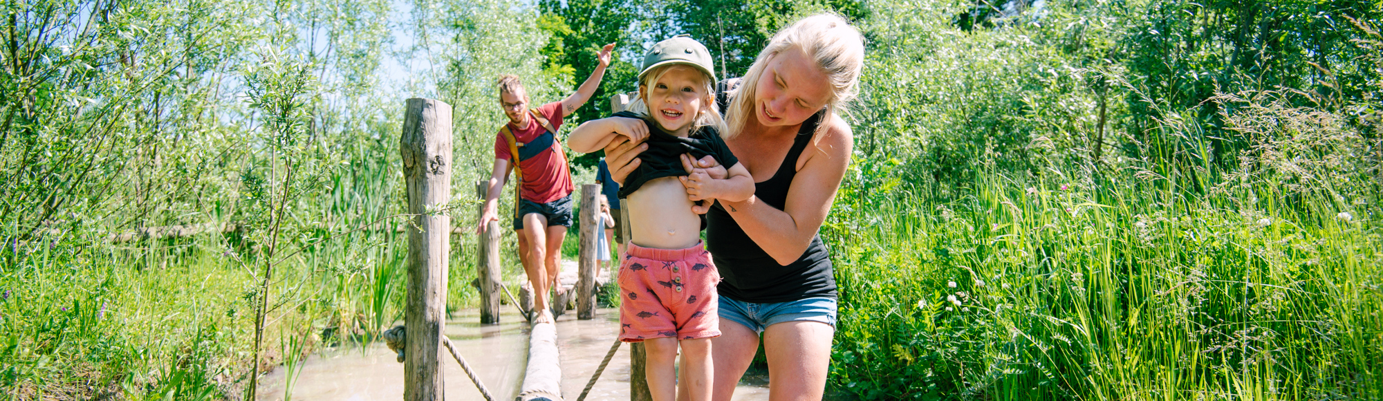 Moeder helpt haar kind met het lopen over de boomstronken in het water bij het blotevoetenpad