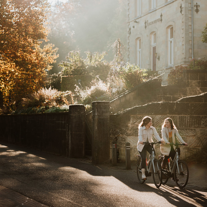 Twee fietsers met op de achtergrond een groot pand van mergel, bomen en een laagstaande zon