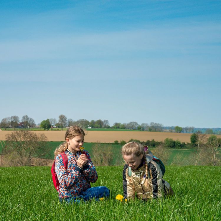 Twee blonde meisjes zitten in het gras en ruiken aan een bloem, met op de achtergrond het limburgse heuvelland. 