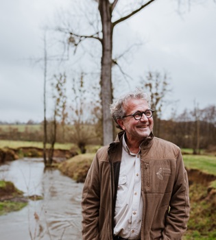 Edmond Staal poseert lachend voor de Geul op een bewolkte dag