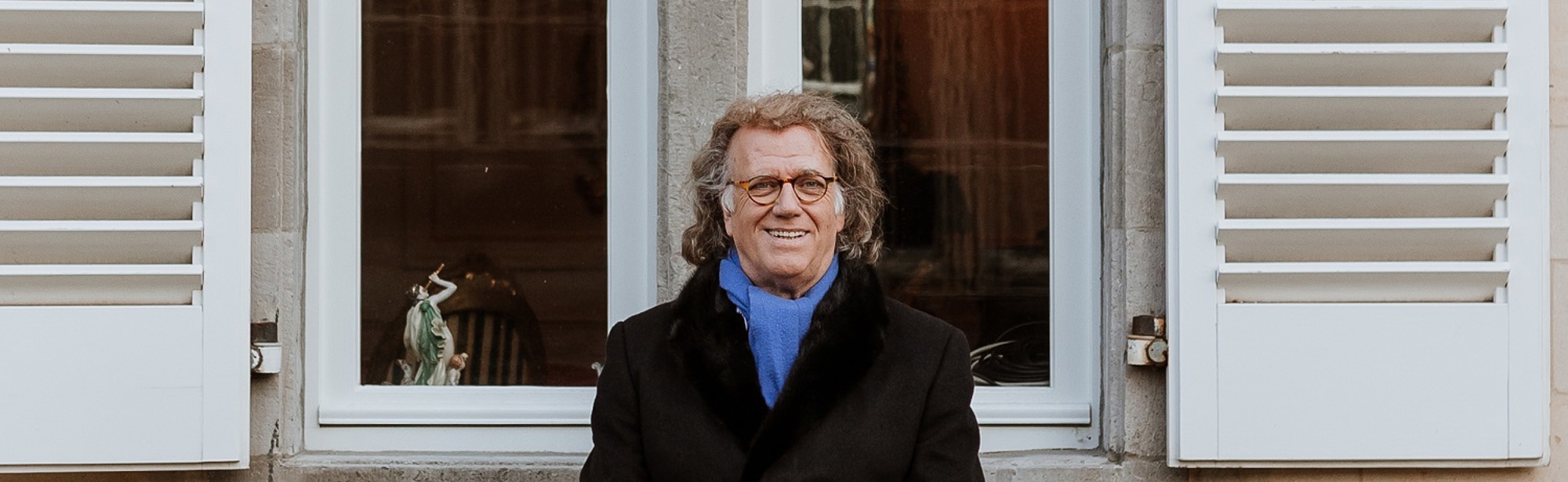 André Rieu poseert op een bankje voor een groot raam met witte luiken