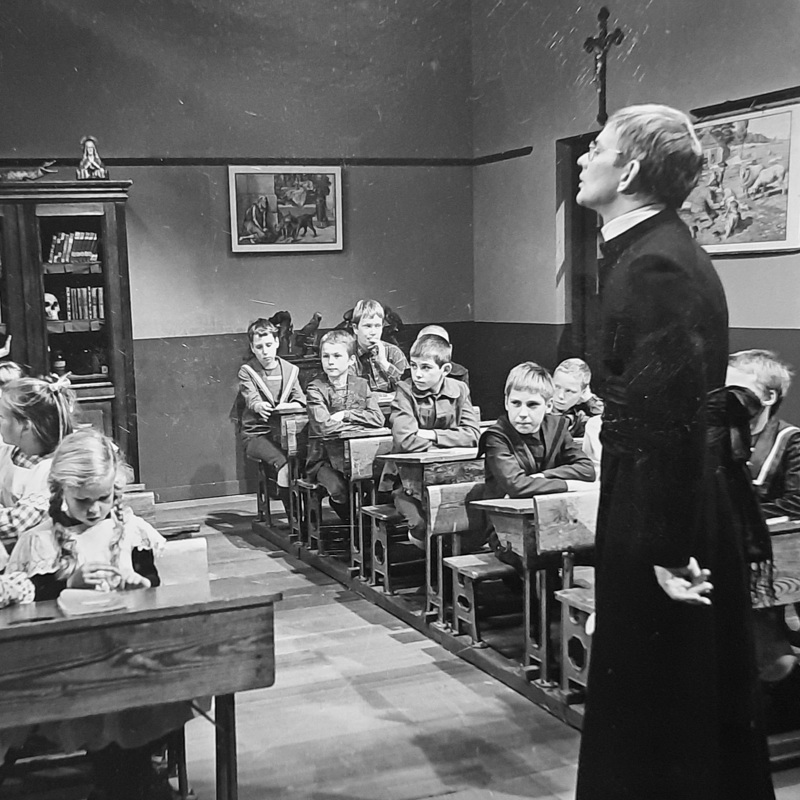 Oude zwart wit foto van Kapelaan Odekerken met kinderen in schoolbankjes in een klaslokaal (Dagboek van een Herdershond)