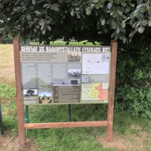 Plaquette met informatie over geheime US Radarinstallatie naast struiken en onder een boom, links staan gekleurde wandelpaaltjes