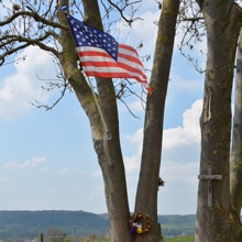 Herdenkingsplaat gemonteerd op een rots tussen twee bomen waarin de Amerikaanse vlag wappert, met uizicht richting Valkenburg