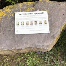 Kleine plaquette op een rots voor een heg met de namen en foto's van zeven omgekomen verzetslieden