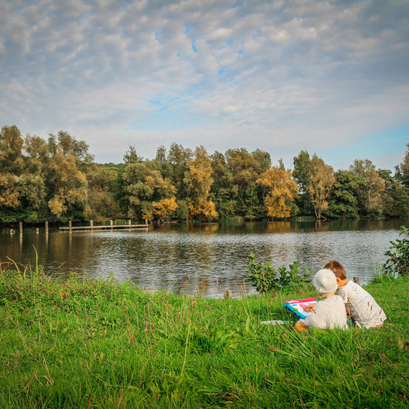 Twee kindjes zitten in het gras bij het water met herfstige bomen op de achtergrond 