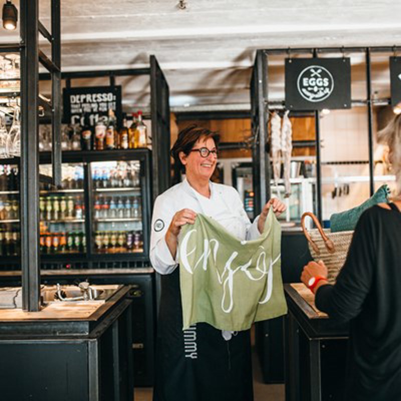 Vrouw in horeca outfit houdt schort vast en lacht naar iemand anders. 