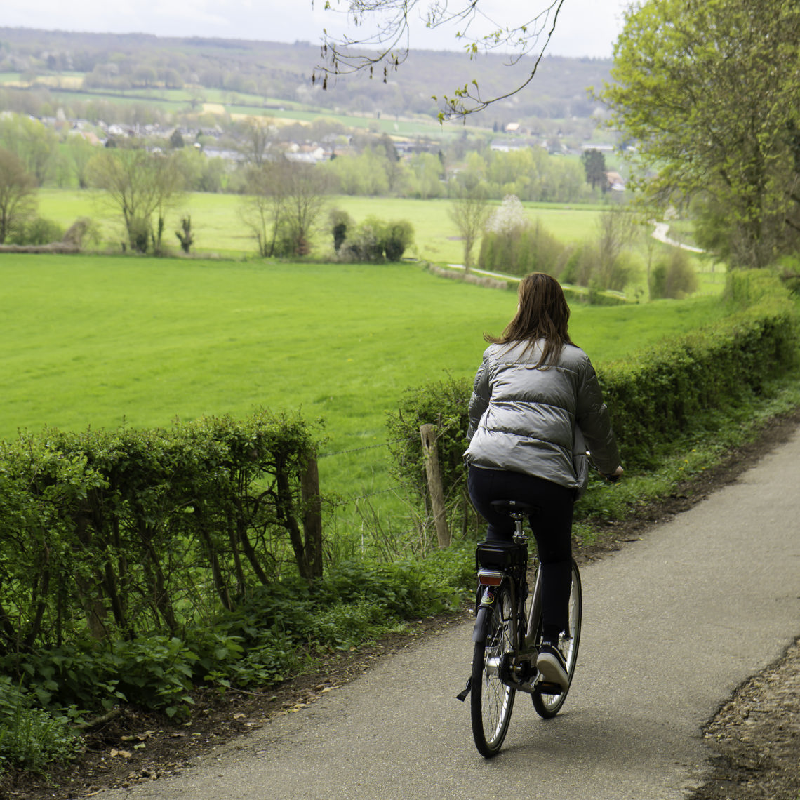 Een vrouw in een zilveren jas fiets over een fietspad met uitzicht over de Limburgse heuvels