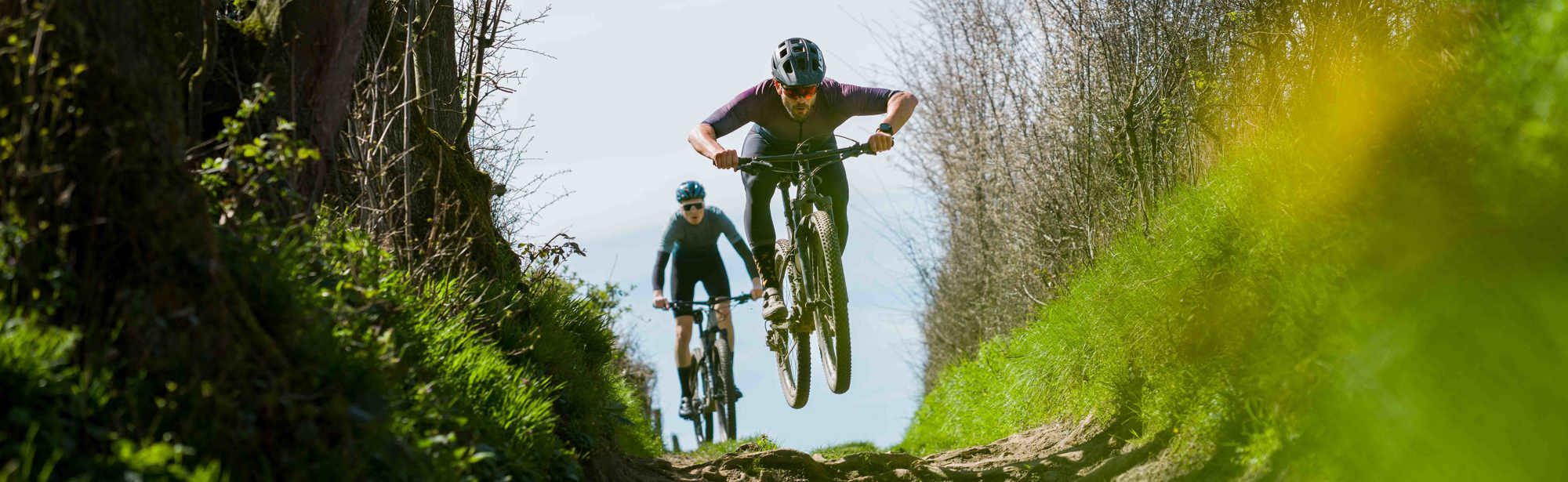 Twee mountainbikers fietsen door een holle weg en een mountainbiker springt in de lucht