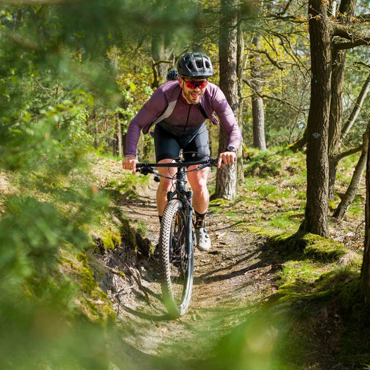 Een mountainbiker fietst lachend over een smal paadje in een zonnig bos