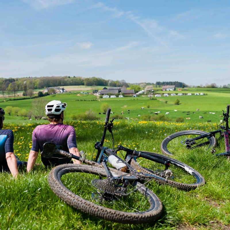 Twee mountainbikers zitten in het gras met hun MTB naast zich en genieten van een prachtig uitzicht over de Zuid-Limburgse heuvels