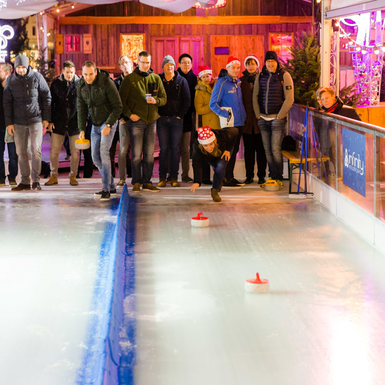 Jonge vrouw met kerstmuts die rode curlingsteen over het ijs schuift bij Wintertijd Heerlen terwijl een groep mensen staat toe te kijken