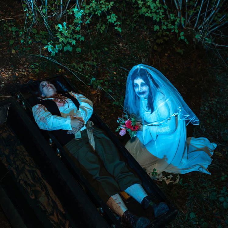 Nachtfoto van bebloed persoon in open doodskist met daarnaast een knielend wit bruidje met wit geschminkt gezicht